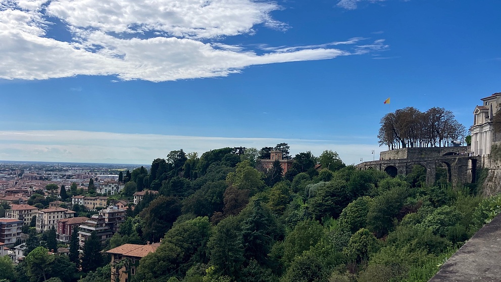 Das Foto zeigt einen Panorama-Blick auf die Stadt Bergamo unter einem strahlend blauen Himmel mit einigen Wolken. Zahlreiche Häuser und grüne Bäume prägen das Stadtbild, rechts ist ein historisches Gebäude zu sehen.