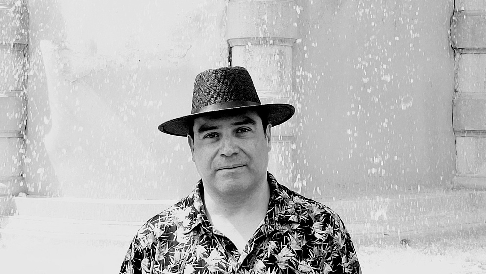 Ein Mann mit einem schwarzen Hut und einem blumigen Hemd steht vor einer sprudelnden Wasserquelle in einem hellen Hintergrund.