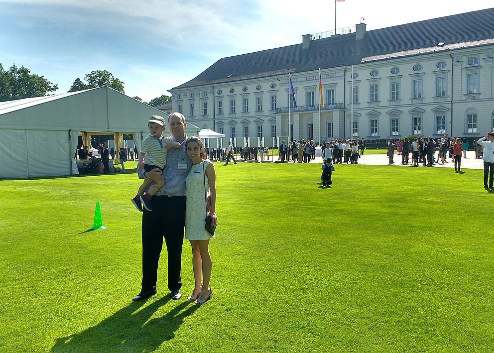Das Foto zeigt Paula Tribelli mit ihrem Mann, der ihren gemeinsamen Sohn auf dem Arm trägt. Das Ehepaar steht auf dem Rasen vor dem Schloss Bellevue und tragen formelle Kleidung.