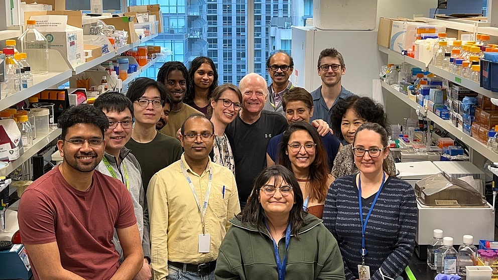 The photo shows a group shot in a University of Toronto science lab with 16 members of the research group, including researchers and professors. All smiling.
