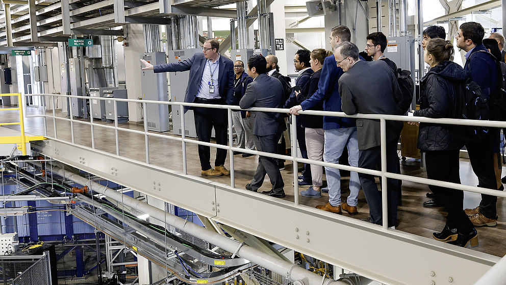 Der Referent zeigt auf eine Maschine, während die Zuhörer auf einer Plattform stehen und interessiert den Erklärungen folgen.