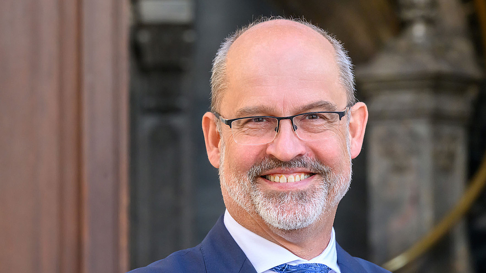 Der Mann mit Brille und einem freundlichen Lächeln steht vor einer eleganten Architektur, gekleidet in einem dunklen Anzug.