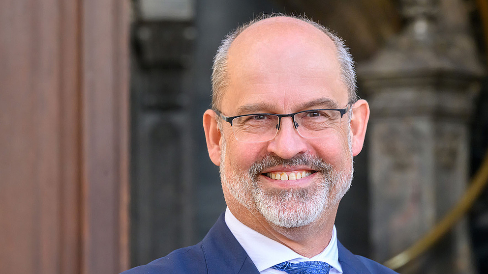 Der Mann mit Brille und einem freundlichen Lächeln steht vor einer eleganten Architektur, gekleidet in einem dunklen Anzug.