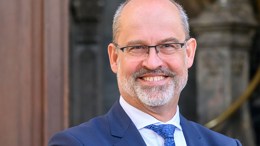 Der Mann mit Brille und einem freundlichen Lächeln steht vor einer eleganten Architektur, gekleidet in einem dunklen Anzug.