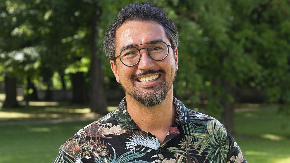 Ein lächelnder Mann mit Brille und einem bunten, tropisches Hemd steht in einem sonnigen Park, umgeben von grünen Bäumen und Rasen.