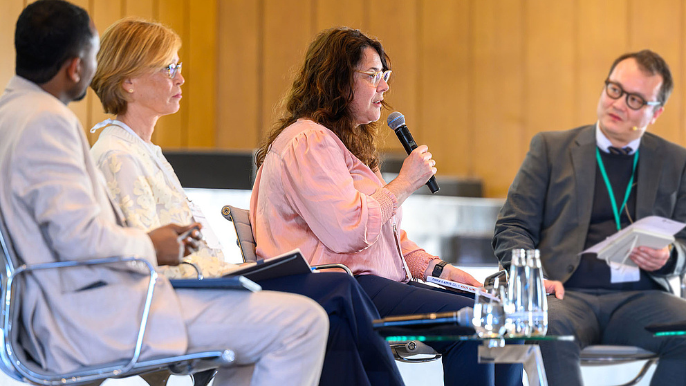 Vier Personen auf Stühlen auf einer Bühne. Eine Frau spricht in ein Mikrophon.