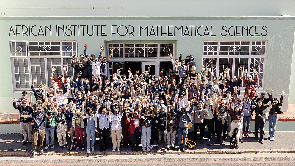 Eine große Gruppe von Menschen steht vor dem African Institute for Mathematical Sciences und hebt die Hände in die Luft.