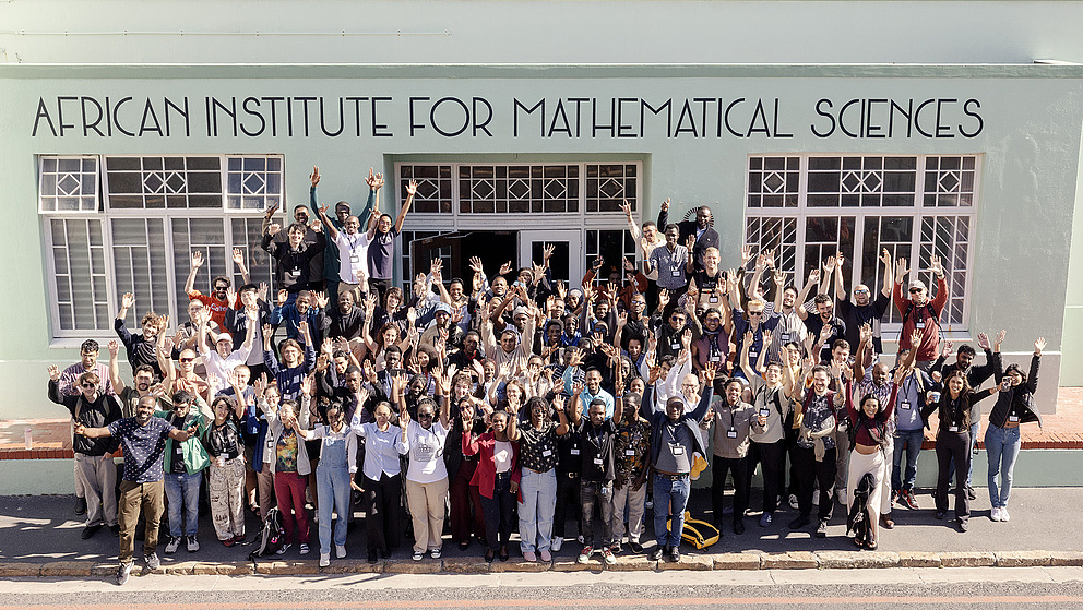 Eine große Gruppe von Menschen steht vor dem African Institute for Mathematical Sciences und hebt die Hände in die Luft.