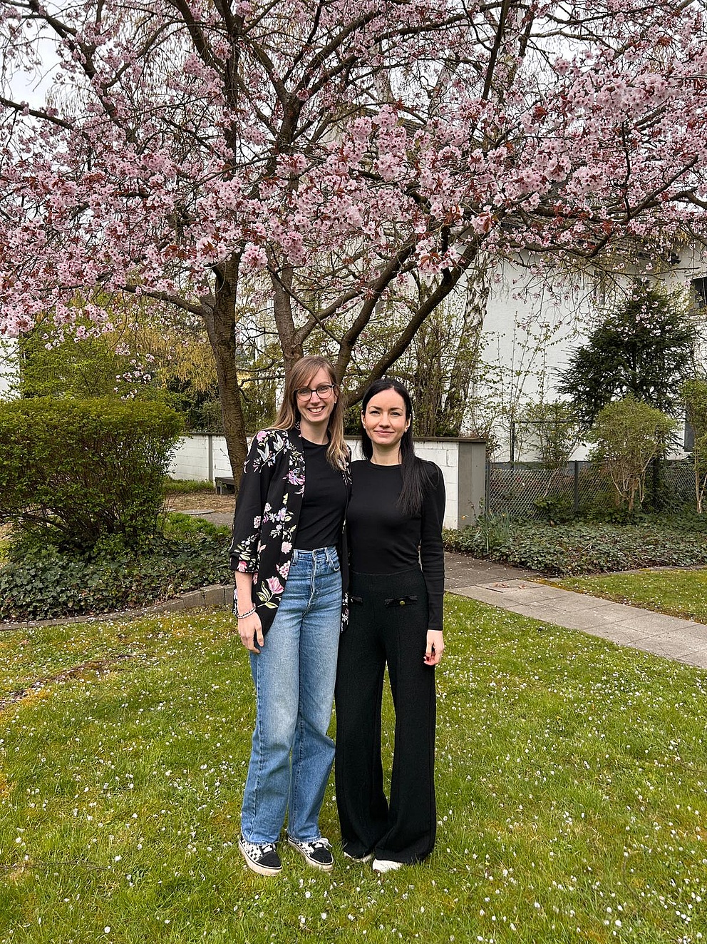 Zwei Frauen stehen lächelnd Arm in Arm im Garten der Humboldt-Stiftung vor einem blühenden Kirschbaum. Die Frau links hat lange blonde Haare, trägt eine Brille, eine geblümte Strickjacke und Jeans. Die Frau rechts hat langes schwarzes Haar und ist schwarz gekleidet. 