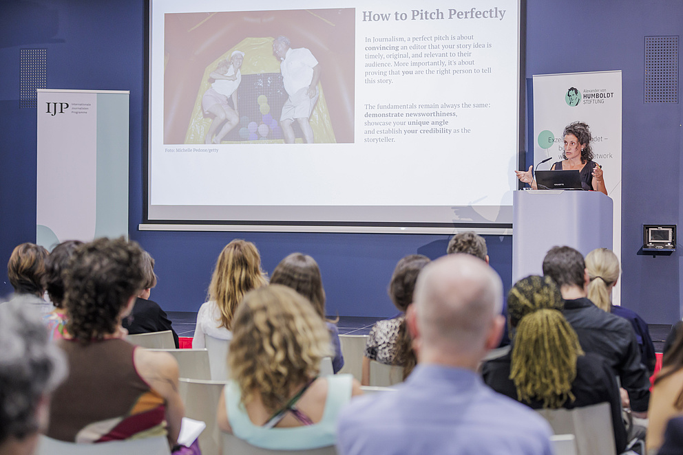 Eine Frau steht auf einer Bühne hinter einem Rednerpult und spricht zum Publikum. Auf der Präsentation im Hintergrund steht "Howto pitch perfectly. In journalism a perfect pitch is about convincing an editor that your story idea is timely, original, and relevant to their audience. More importantly, it's about proving that you are the right person to tell this story. The fundamentals remain always the same: demonstrate newsworthiness, showcase your unique angle and establish your credibility as the storyteller.