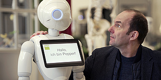 Ein Mensch interagiert mit einem humanoiden Roboter namens Pepper, der eine freundliche Nachricht auf einem Bildschirm zeigt.