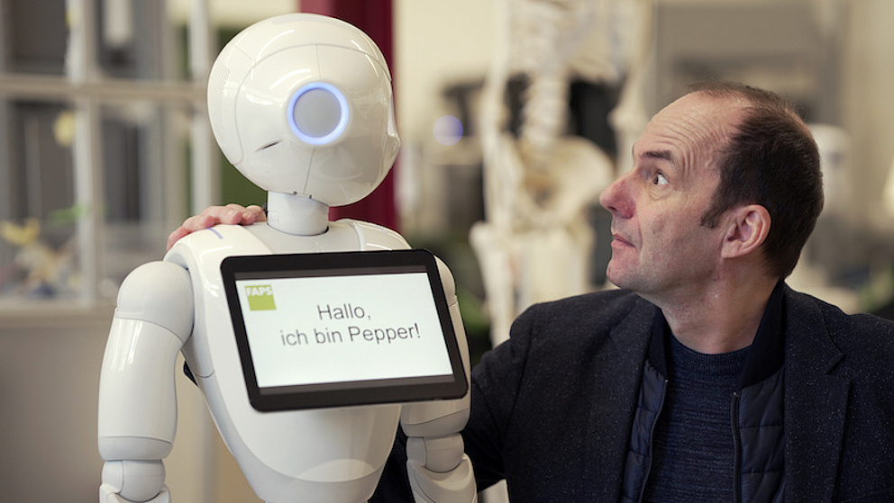 Ein Mensch interagiert mit einem humanoiden Roboter namens Pepper, der eine freundliche Nachricht auf einem Bildschirm zeigt.