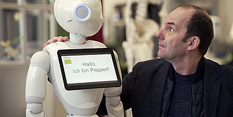 Ein Mensch interagiert mit einem humanoiden Roboter namens Pepper, der eine freundliche Nachricht auf einem Bildschirm zeigt.