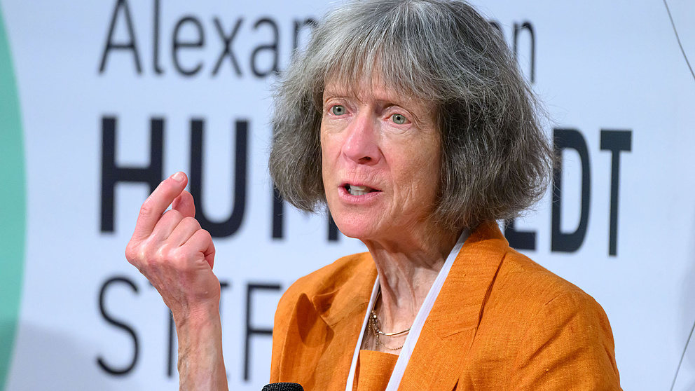 Frau mit orangefarbenem Blazer hält einen Vortrag oder diskutiert vor einem Banner der Humboldt-Stiftung.