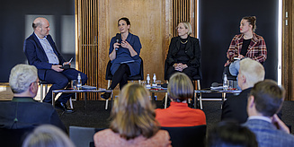 Vier Personen – ein Mann und drei Frauen – sitzen auf Stühlen auf einer Bühne. Eine der Frauen spricht in ein Mikrofon.