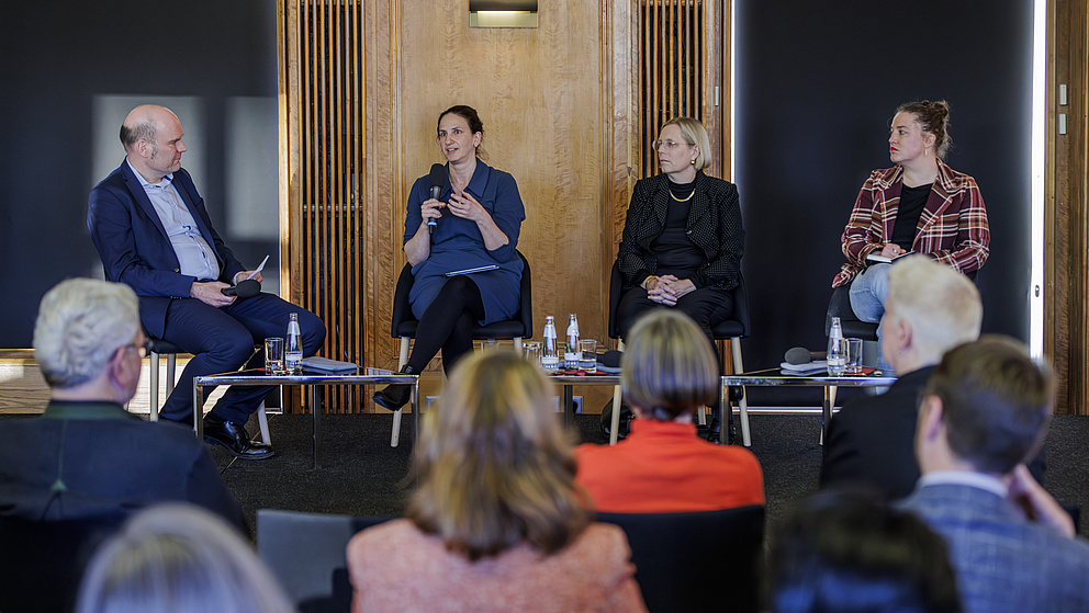Vier Personen – ein Mann und drei Frauen – sitzen auf Stühlen auf einer Bühne. Eine der Frauen spricht in ein Mikrofon.