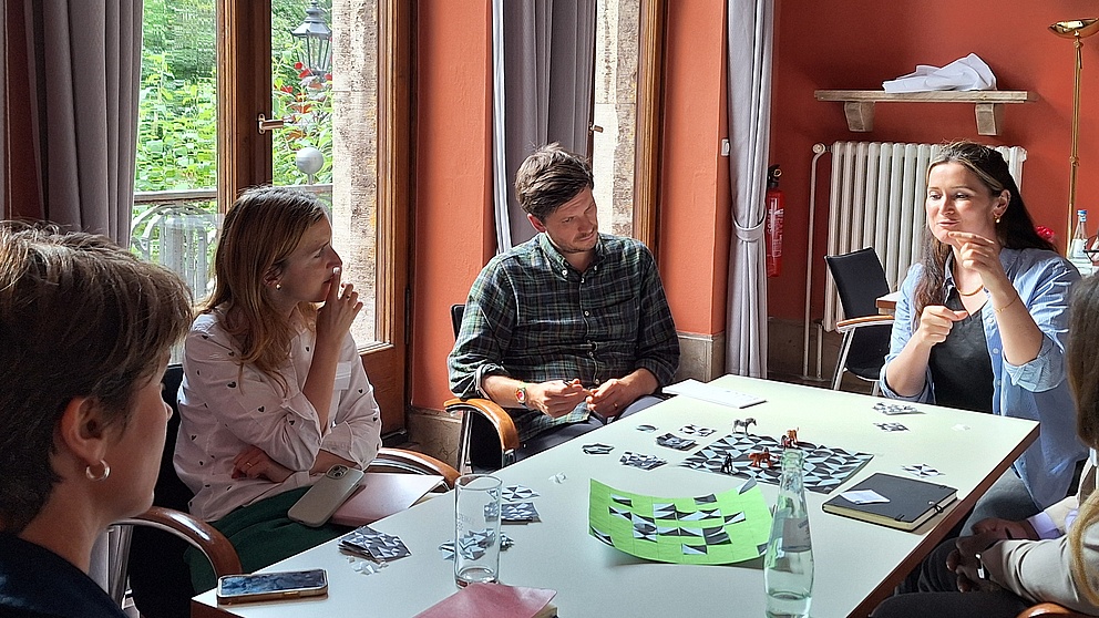 Die Atmosphäre ist entspannt, während eine Frau am Tisch gestikuliert und die anderen Teilnehmer aufmerksam auf ihre Worte reagieren.