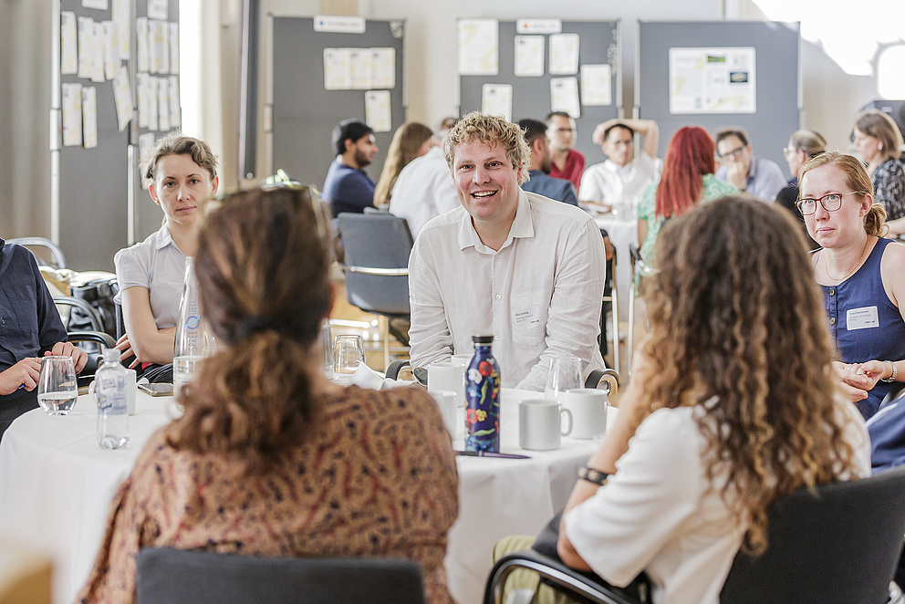 Mehrere Personen sitzen um einen Tisch herum. Im Fokus ist ein Mann, der lacht. 