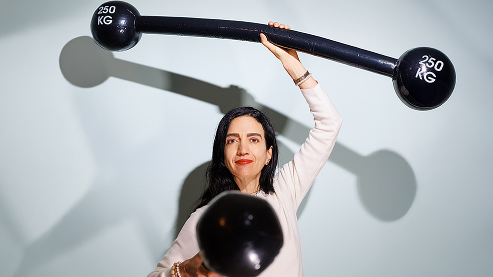 Eine Frau, die zwei Gewichte hochhält, schaut freundlich-bestimmt in die Kamera – ihre Gewichte symbolisieren ihr wissenschaftliches Kernthema „Kinder stärken“.