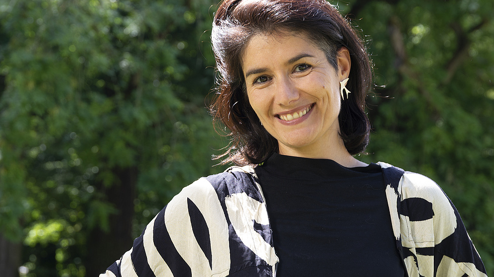 Eine lächelnde Frau mit dunklen Haaren steht in einem sonnigen Park vor grünen Bäumen.