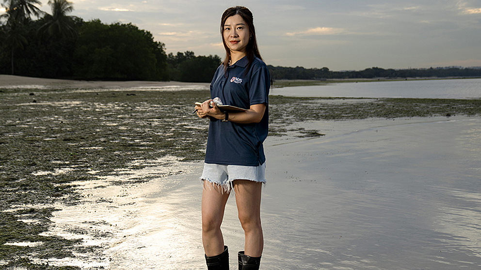 Eine Frau steht im flachen Wasser, trägt Gummistiefel und hält ein Tablet, während sie die Umgebung beobachtet.