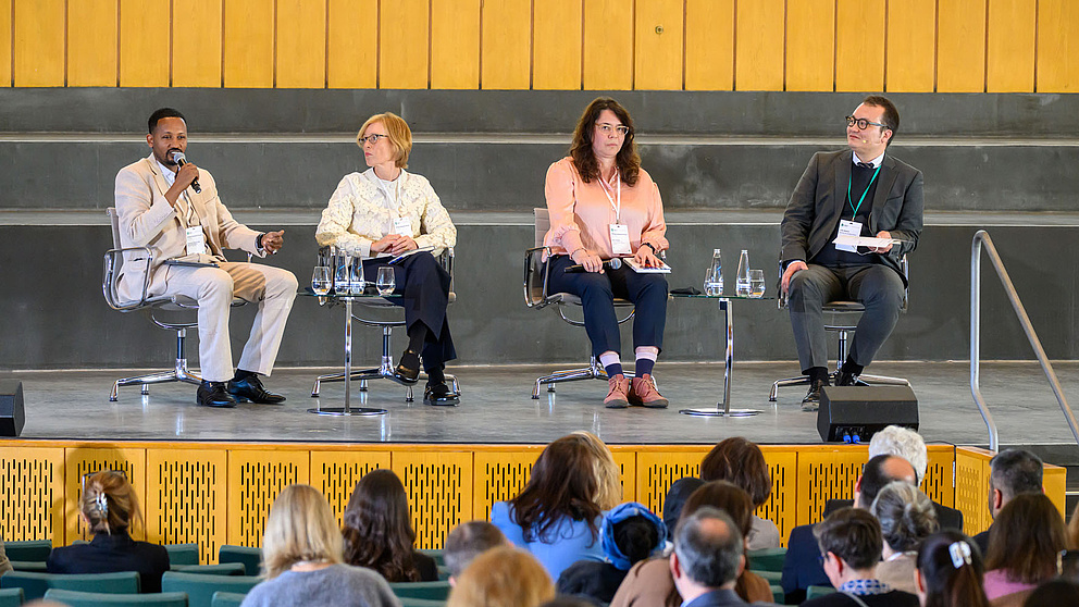 Vier Personen sitzen auf Stühlen auf einer Bühne, einer spricht in ein Mikro.