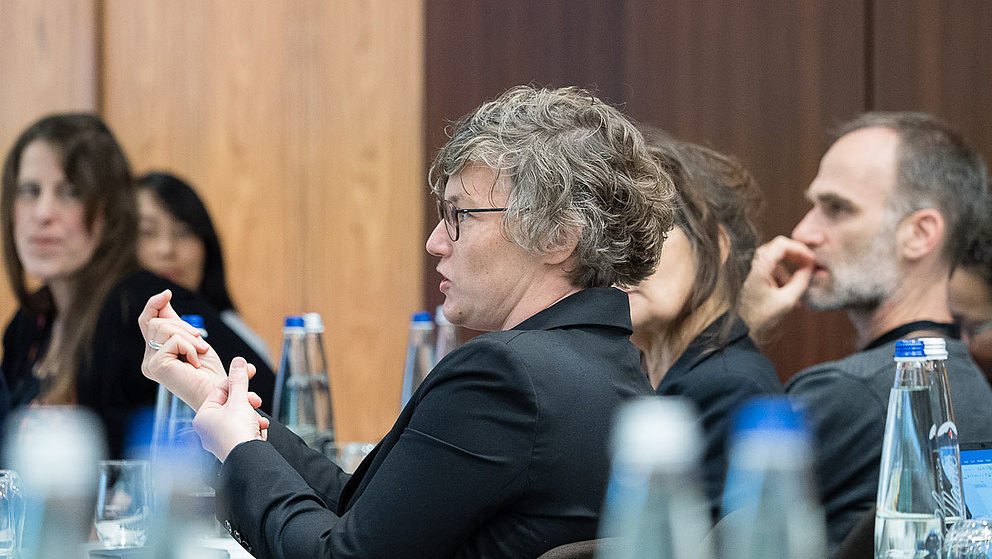 Eine Gruppe von Menschen sitzt an einem Tisch, während eine Person mit Brille nachdenklich gestikuliert und spricht.