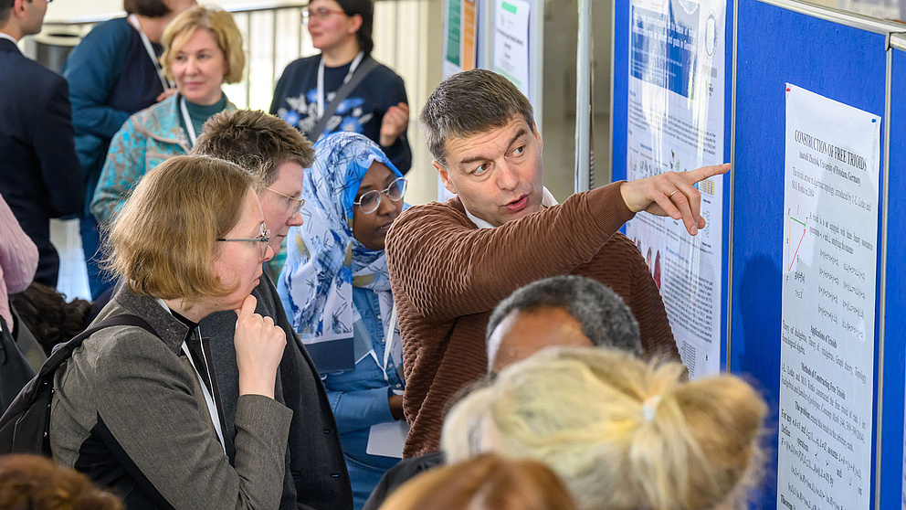 Personen unterhalten sich über ein Poster, ein Mann zeigt darauf. 