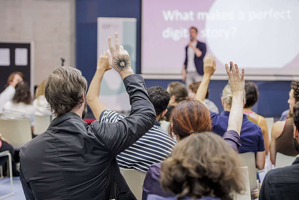 Publikum ist von hinten zu sehen. Ein Mann, der sich meldet, hat ein Tattoo einer schwarzen Sonne auf der Rückhand. 