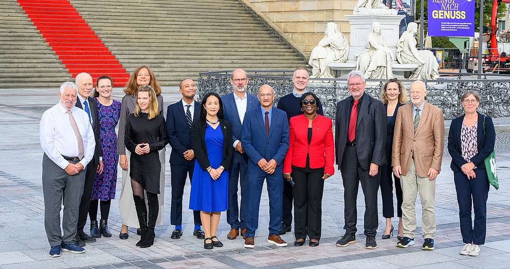 Eine Gruppe von 15 Personen steht vor einer großen Steintreppe mit rotem Teppich und historischen Statuen im Hintergrund.