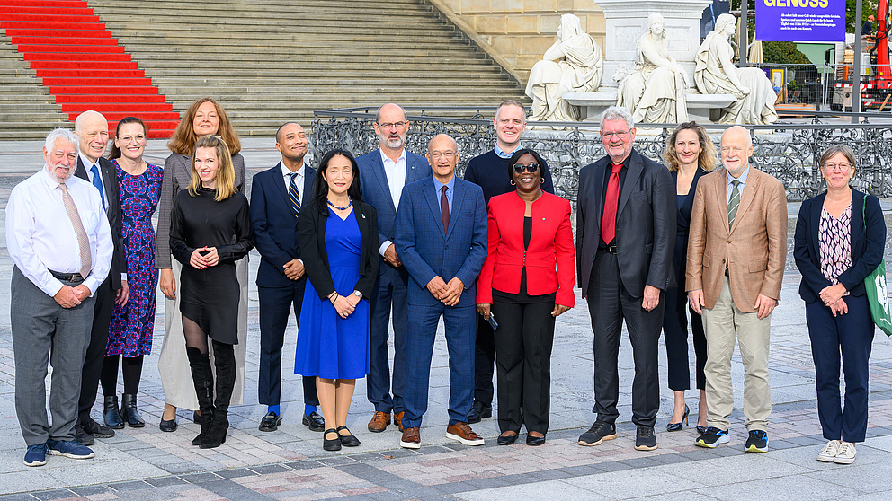 Eine Gruppe von 15 Personen steht vor einer großen Steintreppe mit rotem Teppich und historischen Statuen im Hintergrund.