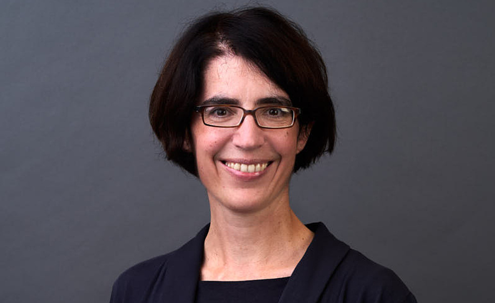 Eine Frau mit kurzen, dunklen Haaren und Brille lächelt freundlich vor einem grauen Hintergrund.