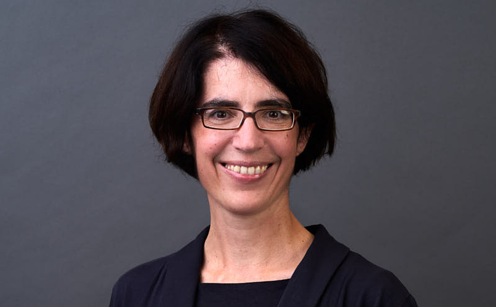 Eine Frau mit kurzen, dunklen Haaren und Brille lächelt freundlich vor einem grauen Hintergrund.