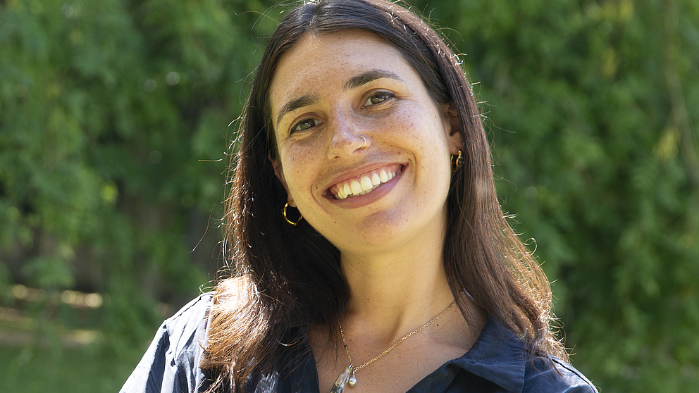 Eine lächelnde junge Frau mit langen, braunen Haaren steht in einem sonnigen Park vor grünen Bäumen.