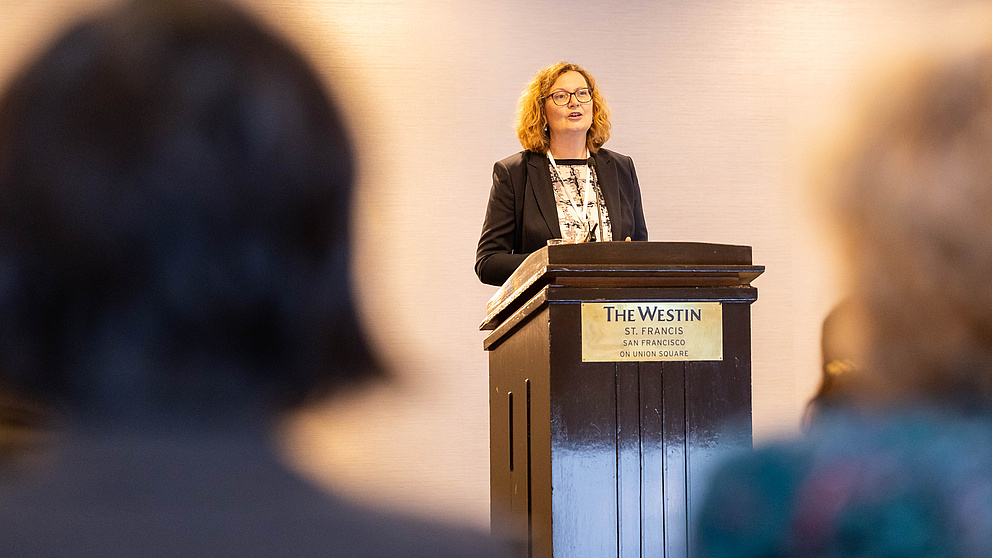 Eine Frau auf einer Bühne hinter einem Rednerpult spricht zum Publikum.