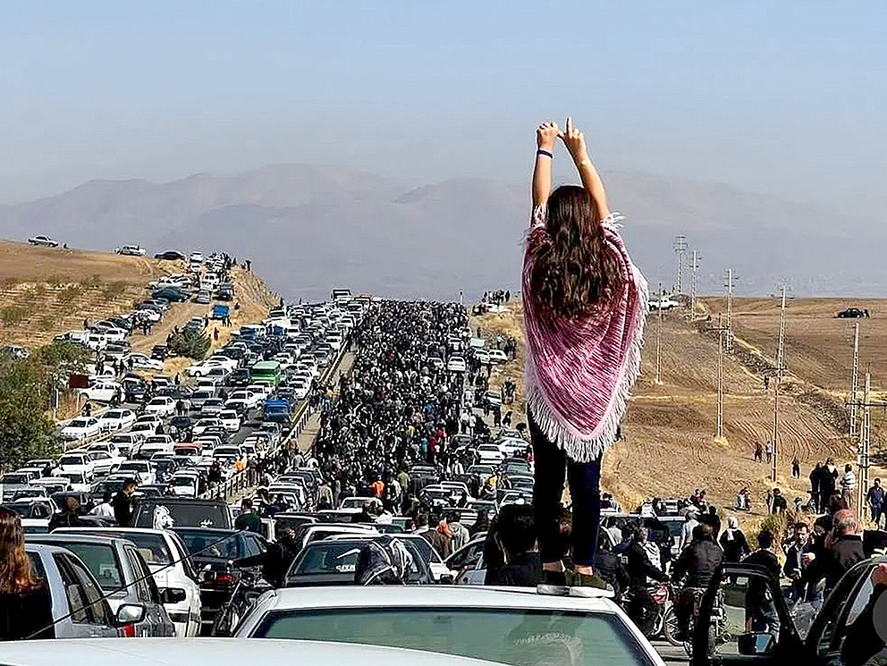 Ein Mädchen steht auf einem Auto und hebt die Hände in die Luft, während eine große Menschenmenge und viele Autos zu sehen sind.