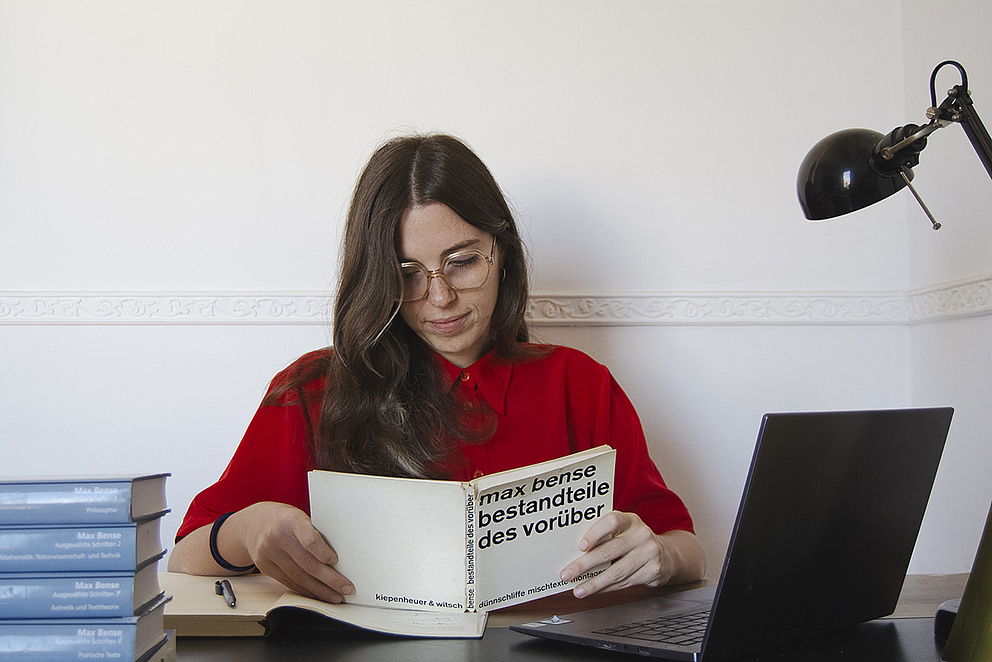 Rosa Coppola, eine junge Frau mit langen braunen Haaren, die eine Brille und ein rotes Hemd trägt, sitzt an einem Schreibtisch. Sie liest ein Buch mit dem Titel „Max Bense – Bestandteile des Vorüber“, neben ihr stehen ein Laptop und ein Stapel Bücher. In der Ecke ist eine schwarze Schreibtischlampe zu sehen.
