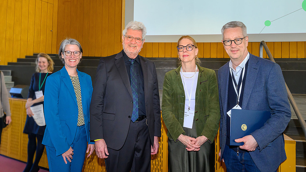 Vier Personen stehen vor einer Bühne und lächeln in die Kamera.