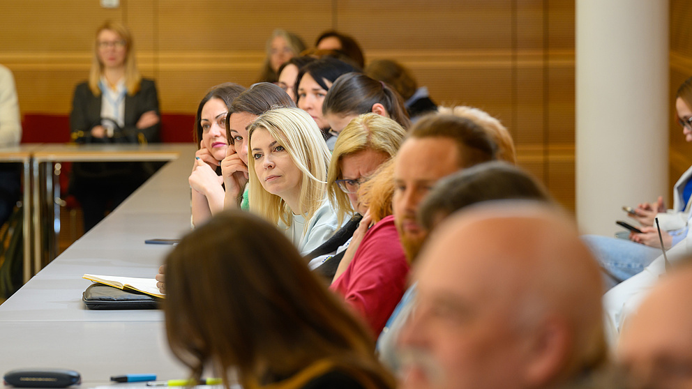 Eine Gruppe von Menschen sitzt in einem Seminarraum und hört zu. Eine blonde aufmerksam zuhörende Frau ist im Fokus.