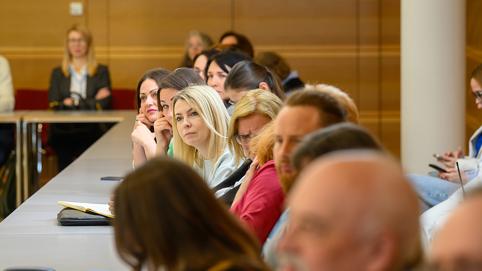 Eine Gruppe von Menschen sitzt in einem Seminarraum und hört zu. Eine blonde aufmerksam zuhörende Frau ist im Fokus.