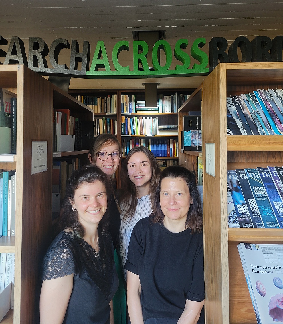 Vier Frauen stehen lächelnd zwischen Bücherregalen in einer Bibliothek. Sie tragen sommerliche Kleidung. Über ihnen hängt ein Holzschriftzug mit der Aufschrift „Research Across Borders“.