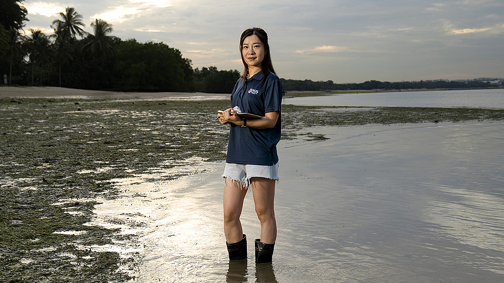 Eine Frau steht im flachen Wasser, trägt Gummistiefel und hält ein Tablet, während sie die Umgebung beobachtet.