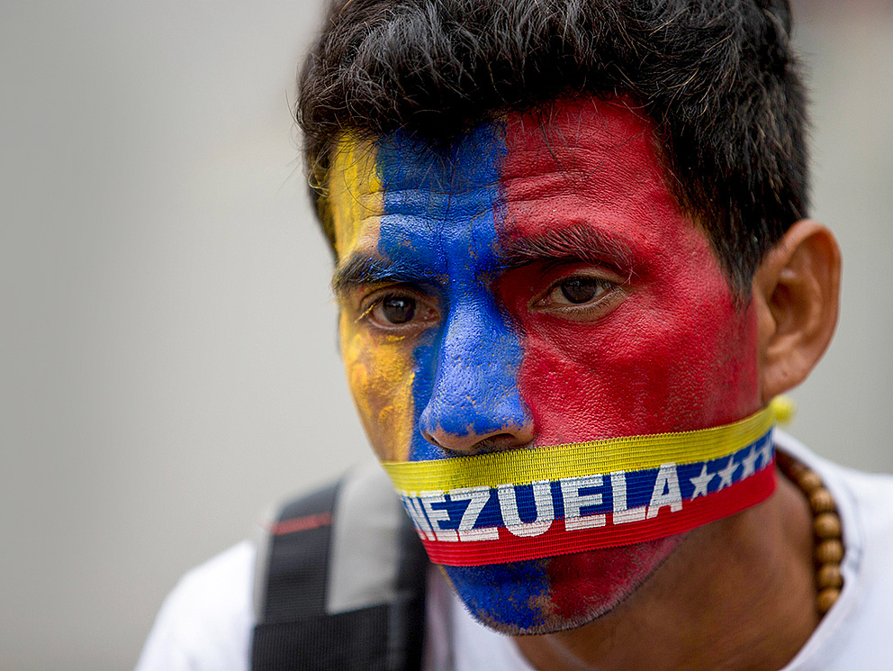 Ein Mann mit rot-blauer Gesichtsbemalung und einem Band mit der Aufschrift 'VENEZUELA' um den Mund, zeigt Protest und Entschlossenheit.