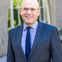 Portrait of a man standing outdoors in front of a blurred building facade with large windows, smiling at the camera. He is wearing rimless glasses, a dark blue suit jacket, a light blue shirt, and a navy tie.