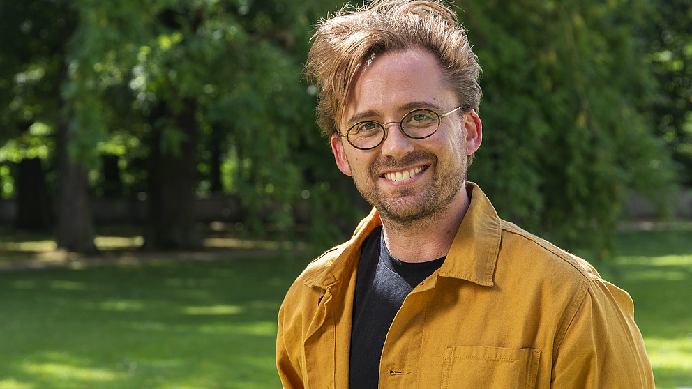 Ein lächelnder Mann mit Brille und gelber Jacke steht in einem sonnigen Park, umgeben von grünen Bäumen und Gras.