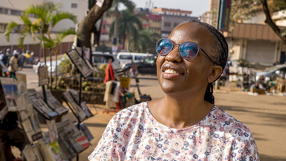 Eine Frau mit Sonnenbrille steht auf einem Platz und blickt in die Sonne, hinter ihr ein Stand mit Zeitungen und Zeitschriften